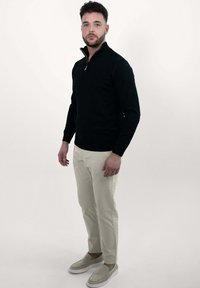 Zwarte pullover met een hoge kraag, beige broek en lichtgrijze instapschoenen. De stof lijkt gebreid met een soepele textuur.