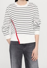 Pull en maille blanche avec des rayures horizontales bleu marine et une couture accent rouge sur le côté gauche. Poignets et ourlet inférieur côtelés. Associé à un jean gris.