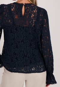 Marineblauw kanten top met bloemmotieven, lange mouwen met geruite manchetten en een sleutelgatachterkant met knoopdetail.