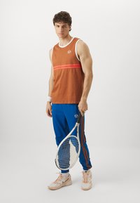 Prunevaba T-särk valge ääre ja punaste triipudega, koos siniste jooksupükste ning oranži külje triibuga. Hoides tennisereketit.
