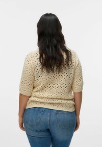 Donna con lunghi capelli scuri che indossa un top corto in crochet beige e jeans blu, di spalle contro uno sfondo bianco uniforme.