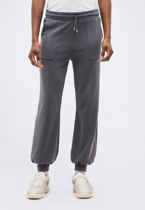 Osoba nosząca ciemnoszare spodnie dresowe z paskiem na sznurek w talii i przednimi kieszeniami, zestawione z jasnobeżowymi sneakersami.