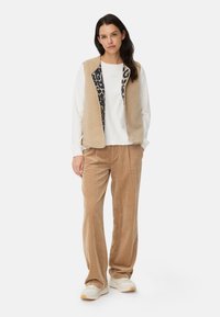 Beige fleece vest with leopard print lining, worn over a white sweatshirt and paired with beige corduroy pants and white sneakers.