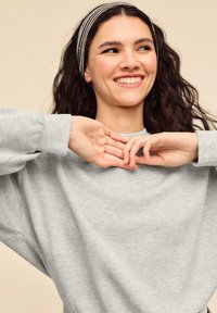 Grauer Sweatshirt mit langen Ärmeln, lockerer Passform. Model trägt ein gestreiftes Stirnband. Weiche Textur, schlichtes Design. Hände nahe am Torso positioniert.