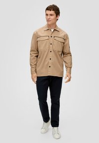Camicia beige con bottoni, due tasche sul petto, con accenti di cuciture nere, colletto, maniche lunghe, abbinata a pantaloni scuri e sneakers bianche.
