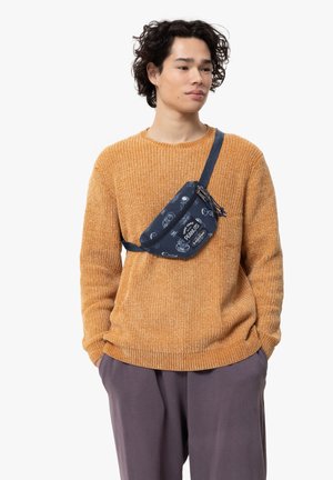 Sac banane bleu à motifs avec fermeture éclair, porté en bandoulière ; pull en laine orange ; pantalon gris ample. Design compact et fonctionnel.