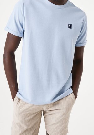 Mężczyzna w jasnoniebieskiej koszulce z krótkim rękawem z małym logo na piersi i beżowych, teksturowanych szortach, z rękami w kieszeniach.