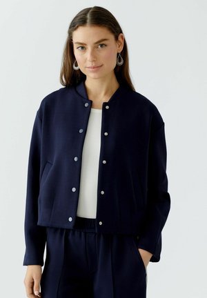Navy cropped jacket with a round collar, snap buttons, and side pockets, made of a smooth fabric. Worn over a white top.