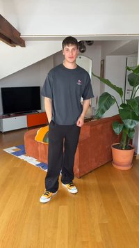 Młody mężczyzna w czarnych spodniach, oversize ciemnoszarej koszulce i żółtych trampkach stoi w salonie z drewnianą podłogą i dużą rośliną doniczkową.