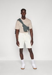 Beiges T-Shirt, weiße Shorts mit Kordelzug und grauer Sweatshirt um die Schultern drapiert. Weiße Sneakers mit hellbraunen Sohlen. Schlichtes Design.
