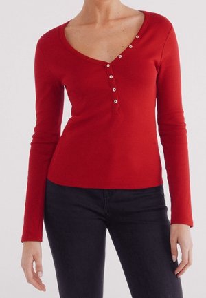 Femme portant un haut rouge moulant à manches longues avec un col asymétrique boutonné et un pantalon noir, debout devant un fond uni.