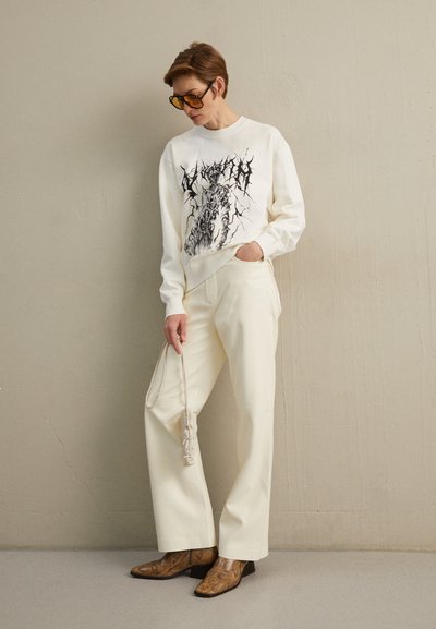 Λευκή μπλούζα με μαύρο γραφικό σχέδιο, κρεμ παντελόνι σε φαρδιά γραμμή και καφέ μποτάκια με σχέδιο φίδι. Αξεσουάρ: ανοιχτόχρωμη τσάντα.