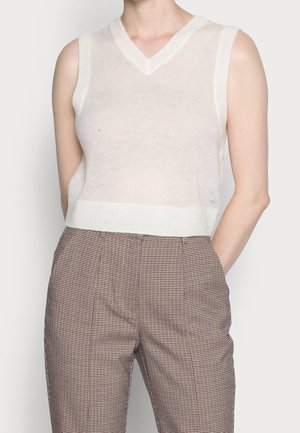 Person trägt ein ärmelloses, weißes Strickoberteil mit V-Ausschnitt, kombiniert mit hochtaillierten, braun-weiß karierten Hosen, die Hände in den Taschen.