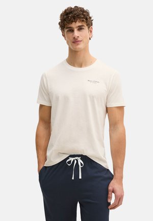 Junger Mann mit lockigem Haar, der ein weißes T-Shirt von Marc O’Polo und marineblaue Hose mit Kordelzug trägt, steht mit einer Hand in der Tasche.