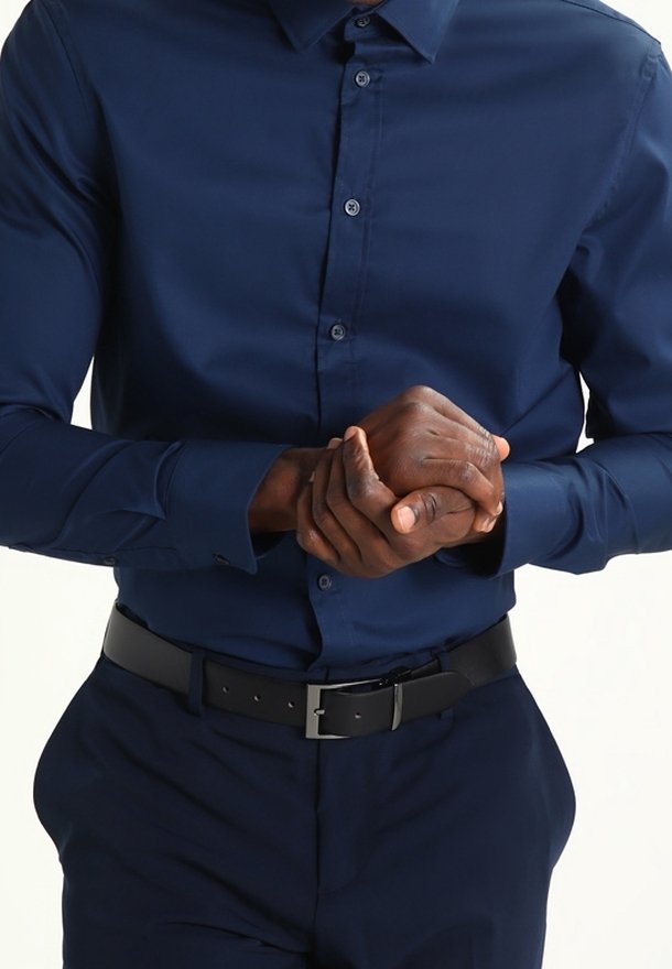 Mężczyzna noszący granatową koszulę zapinaną na guziki i spodnie, składa dłonie przed sobą, prezentując ciemną skórę oraz czarny pasek.