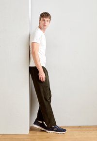 White short-sleeve t-shirt, dark green corduroy pants, and navy sneakers. Man leaning against a wall, with a textured background.
