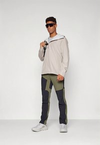 Lys beige vandtæt jakke med hætte, kombineret med mørkegrønne og marineblå lap-patch bukser. Modellen har sorte solbriller og hvide sneakers på.