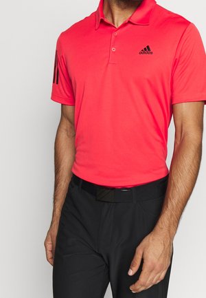Hombre con camisa polo Adidas de color rojo brillante con logo negro y pantalones negros con cinturón negro sobre un fondo claro y liso.