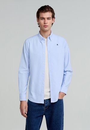 Hellblaues Button-Down-Hemd aus Baumwolle mit einem kleinen schwarzen Totenkopf-Logo auf der Brust, kombiniert mit blauen Jeans.