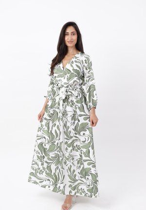 Femme portant une longue robe blanche avec des motifs feuillus verts, debout et regardant vers l'avant sur un fond blanc uni.