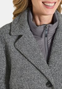 Frau lächelt, trägt einen strukturierten grauen Wollmantel über einer grauen Daunenjacke mit sichtbarem Metallreißverschluss.