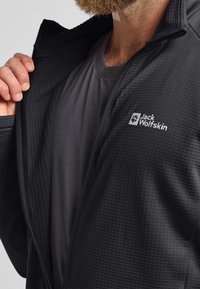 Schwarze gesteppte Jacke mit einem Reißverschluss auf der Vorderseite, darunter ein graues Basisshirt. Logo auf der linken Brustseite. Strukturierter Stoff sichtbar.