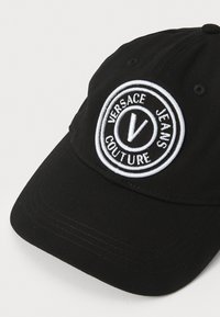 Juodas medvilninis beisbolo kepuraitė su siuvinėtu apvaliu logotipu, kuriame yra tekstas "Versace Jeans Couture" ir iškilia "V" centre.