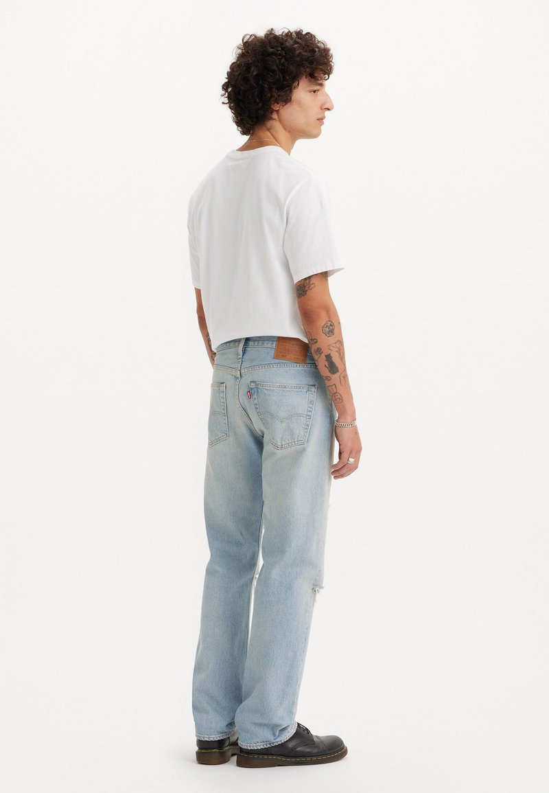 Camicia bianca a maniche corte, jeans scorciati azzurri strappati, scarpe nere. Presenta cuciture in evidenza, una toppa in pelle sui jeans e una vestibilità comoda.
