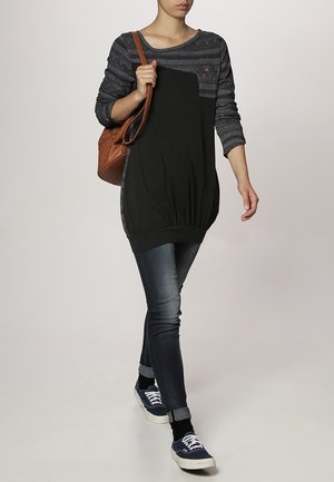 Musta tunika yläosa, jonka harmaissa kuviollisissa hihoissa on kankaan yksityiskohtia, vyötärö kiristettynä, yhdistettynä tummiin skinny-farkkuihin ja tummansinisiin lenkkikenkiin, pitäen ruskeaa käsilaukkua.