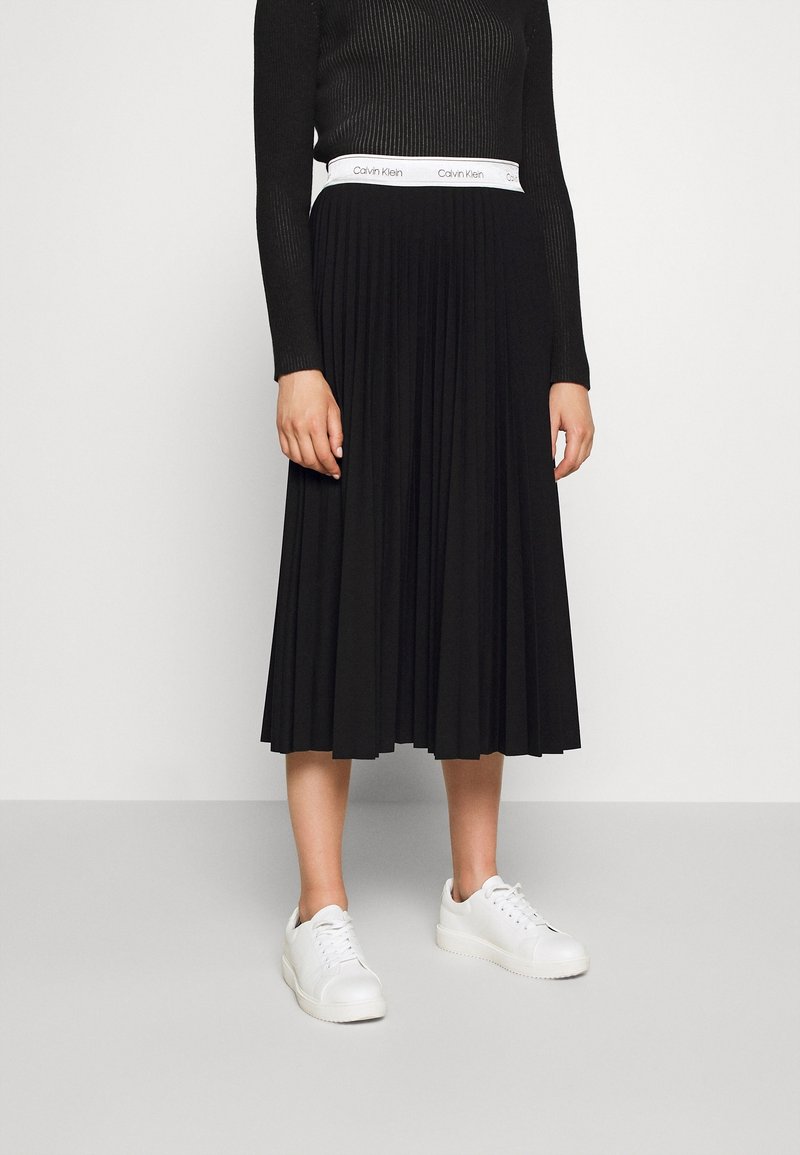 Femme portant une jupe midi plissée noire avec une ceinture blanche Calvin Klein, un haut noir à manches longues côtelé et des baskets blanches.