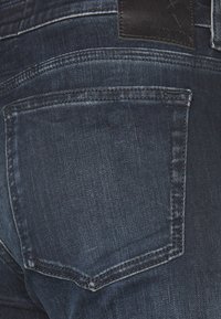 Vaqueros de mezclilla oscura con un bolsillo trasero, que cuentan con costuras blancas y un parche de marca de cuero. La tela parece ligeramente texturizada y desgastada.