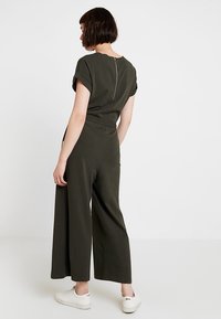Mujer con cabello corto y oscuro, vestida con un mono verde oliva con cremallera trasera y zapatillas blancas, de pie frente a un fondo blanco.