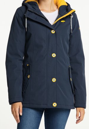 Woman wearing navy blue jacket with yellow lining, yellow buttons, and hood with drawstrings, paired with blue jeans.