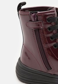 Burgundy patent leather boot with black laces, side zipper, and textured rubber sole. Features stitched accents and a pull tab at the top.
