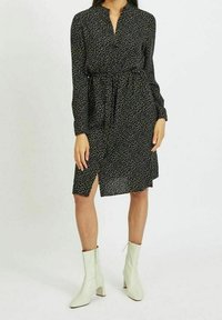 Žena v černých dlouhých šatech s bílým tečkovaným vzorem, s předním rozparkem a bílými kotníkovými botami, stojící na neutrálním pozadí.