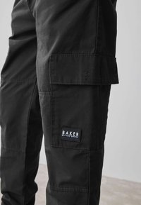 Melni kravas bikses izturīgā audumā, ar sānu kabatu un etiķeti "BAKER by TED BAKER". Dizainā iekļautas redzamas vīles un akcenti.