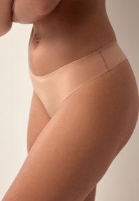 Gros plan d'une personne portant un sous-vêtement beige sans couture, montrant le côté de la taille, de la hanche et du haut de la cuisse sur un fond neutre.
