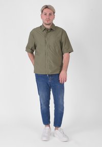 Maglietta verde oliva a maniche corte con taschino frontale, abbinata a jeans slim-fit blu e sneaker bianche, in piedi su uno sfondo semplice.