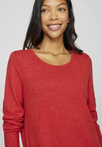 Pull en maille rouge avec un col rond et des manches longues, présentant une texture douce et une coupe décontractée.