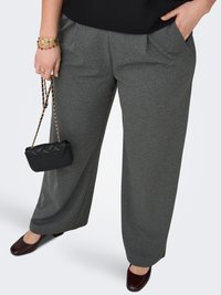 Vrouw draagt grijze broek met wijd uitlopende pijpen, donkerrode schoenen, zwart topje, en houdt een kleine zwarte doorgestikte tas met kettingband en gouden armbanden vast.