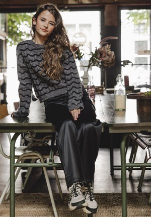 Grijze trui met golvend zwart patroon, zwarte satijnen broek en sneakers met patroon. Het model zit op een houten tafel in een cafésetting.