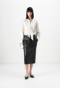 Kvinne iført hvit bluse med sløyfe, svart skinn midi skjørt, svart hæl sko, holder en hvit clutch, står mot en ensfarget hvit bakgrunn.