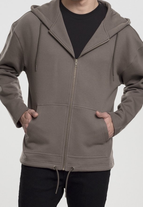 Persona che indossa una felpa grigia con zip e cordini, con le mani nelle tasche anteriori, e pantaloni neri su uno sfondo semplice.