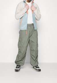 Giacca in pile di colore azzurro chiaro e crema con dettagli arancioni; abbinata a pantaloni cargo larghi di colore verde oliva; sneakers bianche e nere.