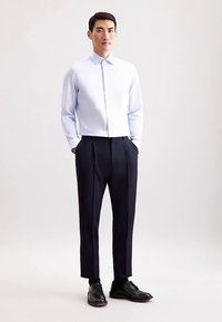 Lichtblauw gestreept overhemd met kraag, gedragen met donkere geplooide broek en zwarte schoenen, staand tegen een neutrale achtergrond.