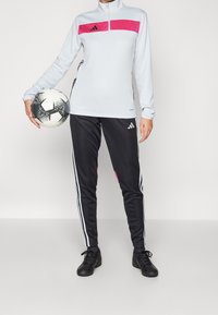 Světlá šedá dlouhá sportovní mikina s růžovým horizontálním pruhem a černé teplákové kalhoty s bílými pruhy. Drží černobílý fotbalový míč.