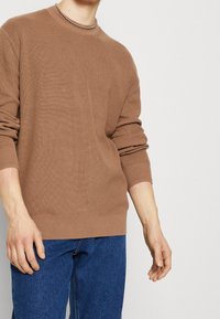 Brązowy sweter prążkowany z okrągłym dekoltem i długimi rękawami, wyróżniający się teksturowanym materiałem i luźnym krojem, połączony z niebieskimi dżinsami.