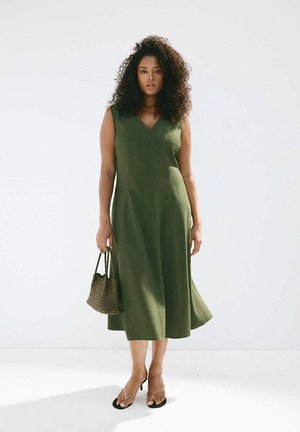 Mujer con cabello rizado que lleva un vestido midi verde sin mangas, sostiene un bolso marrón tejido y lleva sandalias negras con tacón.