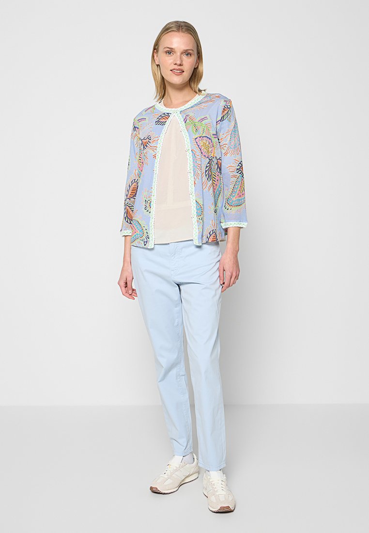 Hellblauer Strickjacke mit langen Ärmeln und bunten Blattmustern; kombiniert mit einem cremefarbenen Oberteil und hellblauen Hosen. Weiße Turnschuhe vervollständigen das Outfit.