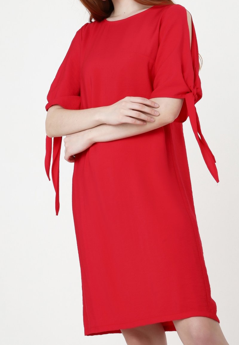 Robe rouge mi-longue à manches échancrées au coude nouées, encolure ronde, portée par une personne aux bras croisés.